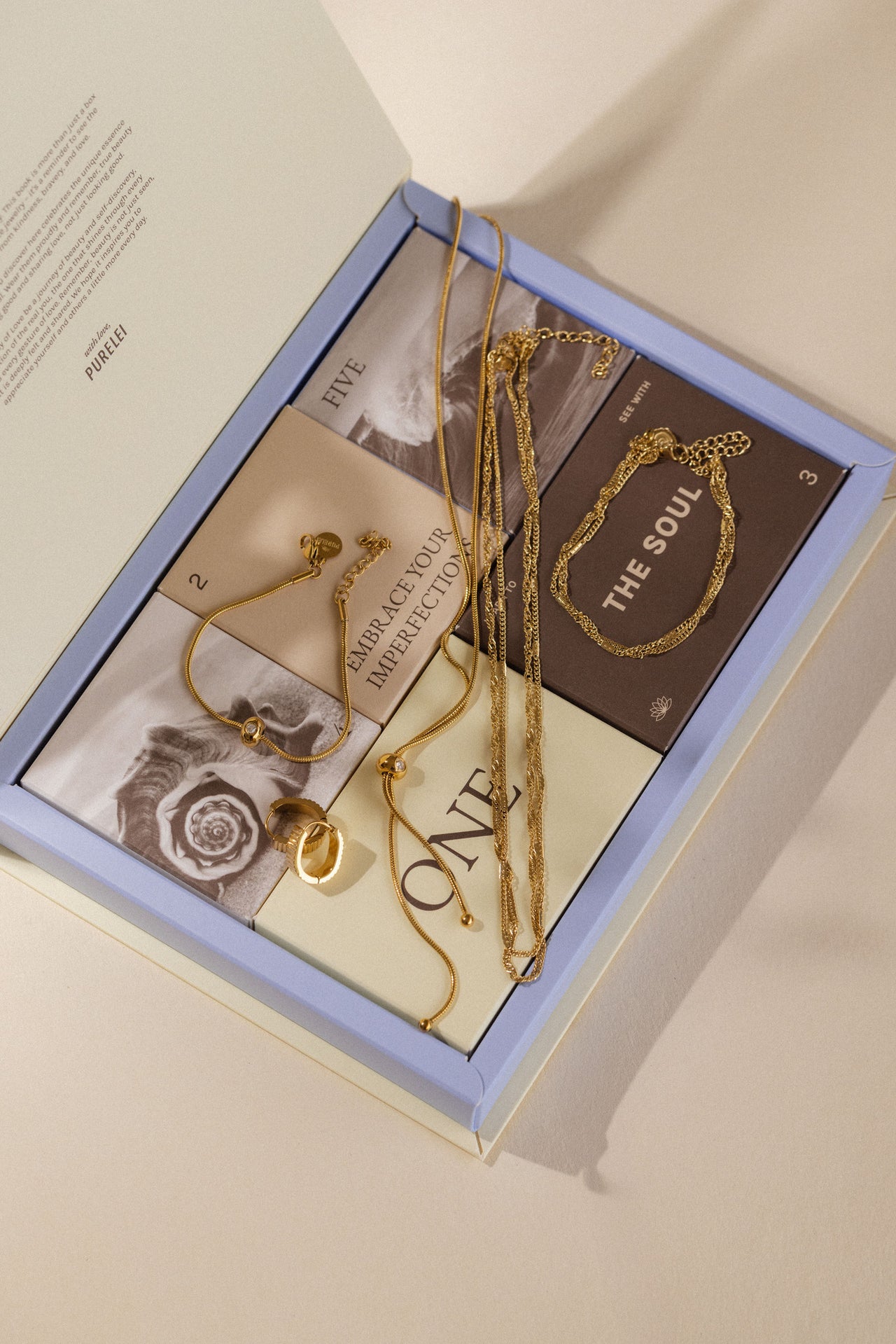Goldene Schmuckstücke in einer eleganten Box von PURELEI, umgeben von inspirierenden Karten.