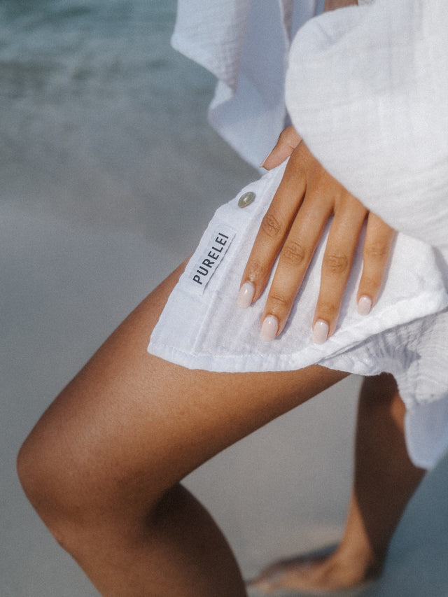 Weißes Tuch mit PURELEI-Label, getragen am Strand, umgeben von sanften Wellen.
