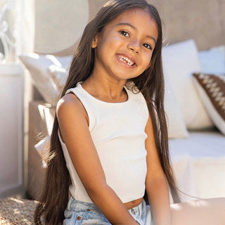 Lächelndes Mädchen in weißem Oberteil, PURELEI inspiriert, mit langen Haaren in heller, freundlicher Umgebung.