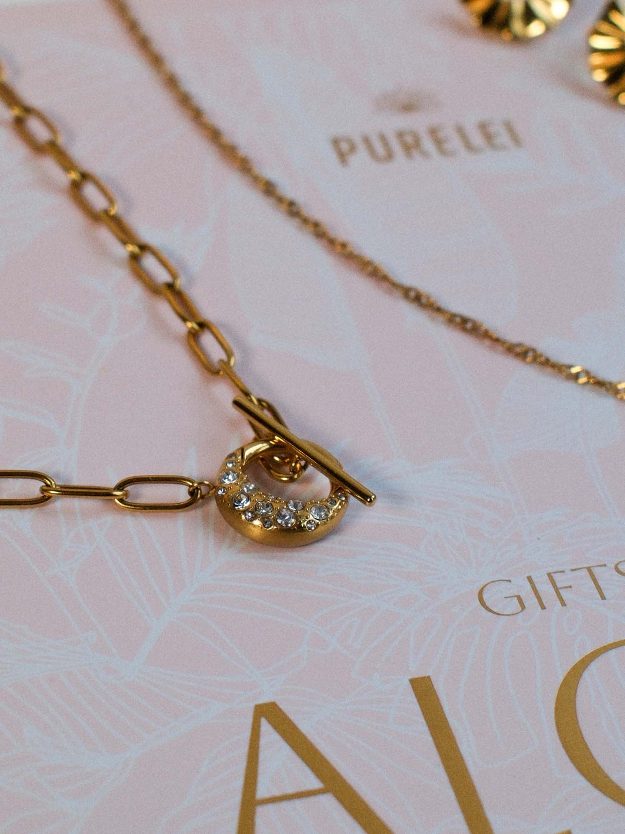 PURELEI Air Schmuckset mit vergoldeter Kette, Kristallanhänger und feinem Toggle-Verschluss, elegant auf Pastellhintergrund.