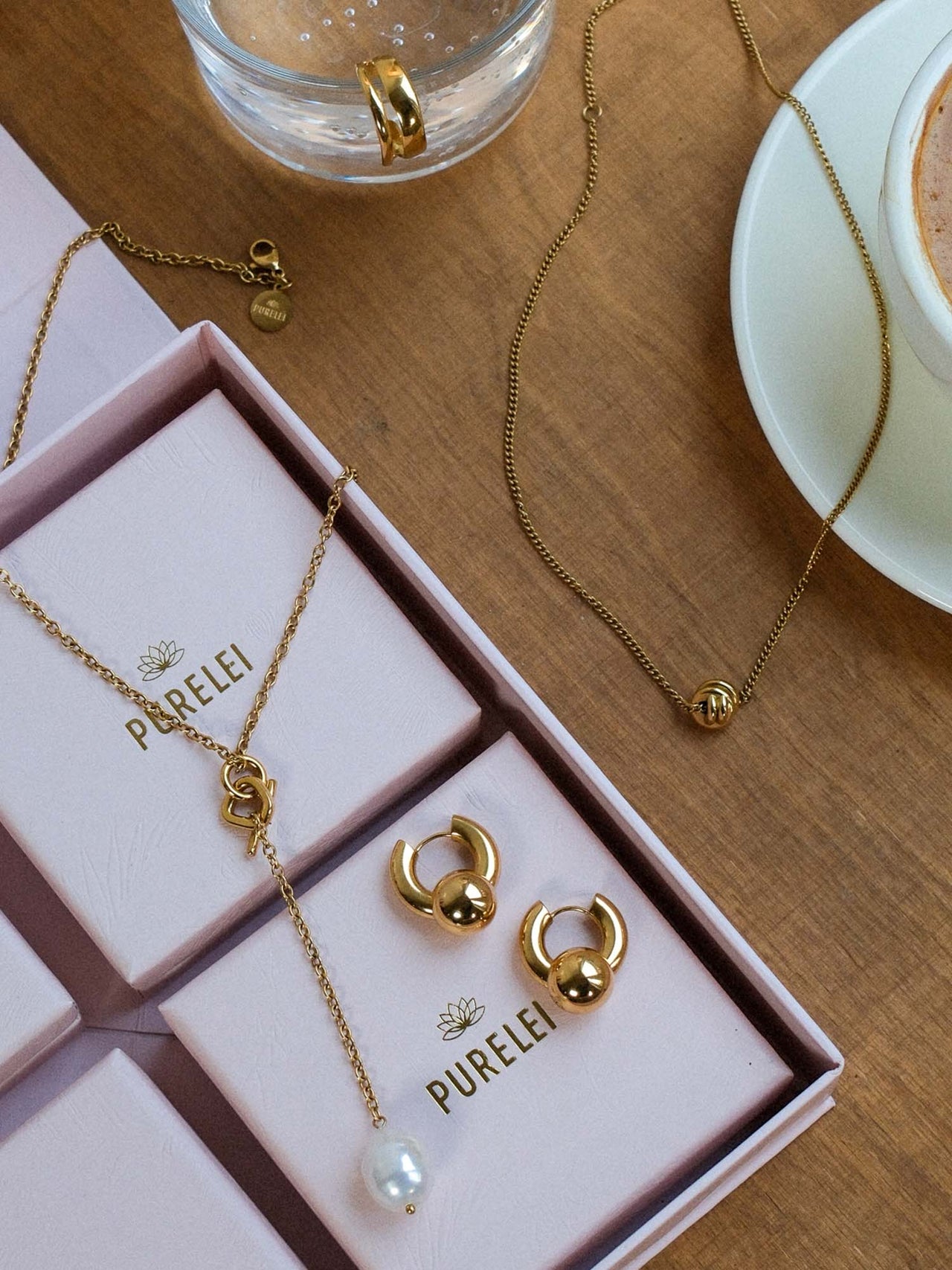PURELEI Aloha Schmuckset mit goldfarbenen Ketten, Perlenanhänger und wandelbaren Creolen, elegant auf Holztisch inszeniert