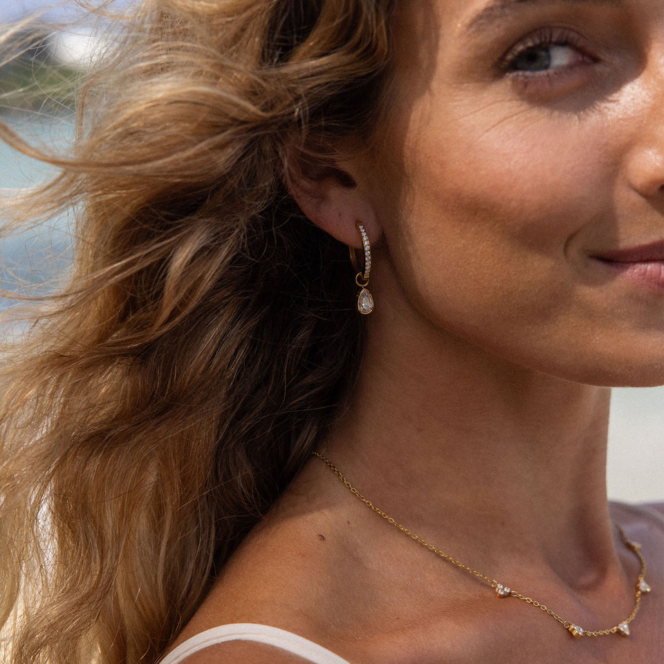 Felice Ohrringe von PURELEI, goldene Creolen mit weißem Zirkonia-Anhänger, elegant und feminin am Strand getragen