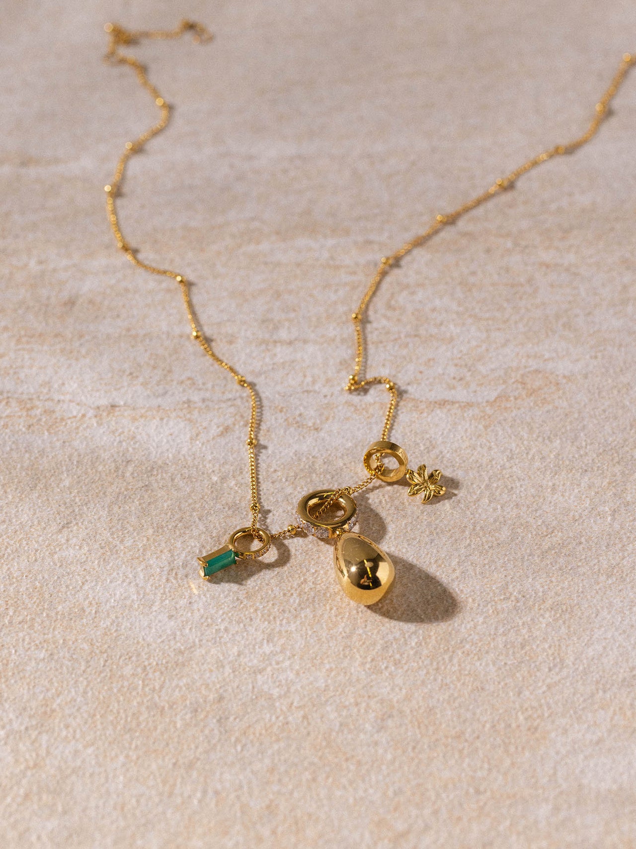 Li’ili’i Kette von PURELEI, vergoldet, filigran mit Tropfen- und Blumenanhänger, elegant auf sandfarbenem Untergrund.