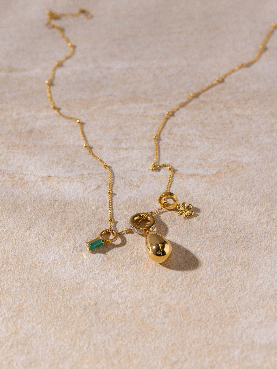 Li’ili’i Kette von PURELEI, vergoldet, filigran mit Tropfen- und Blumenanhänger, elegant auf sandfarbenem Untergrund.