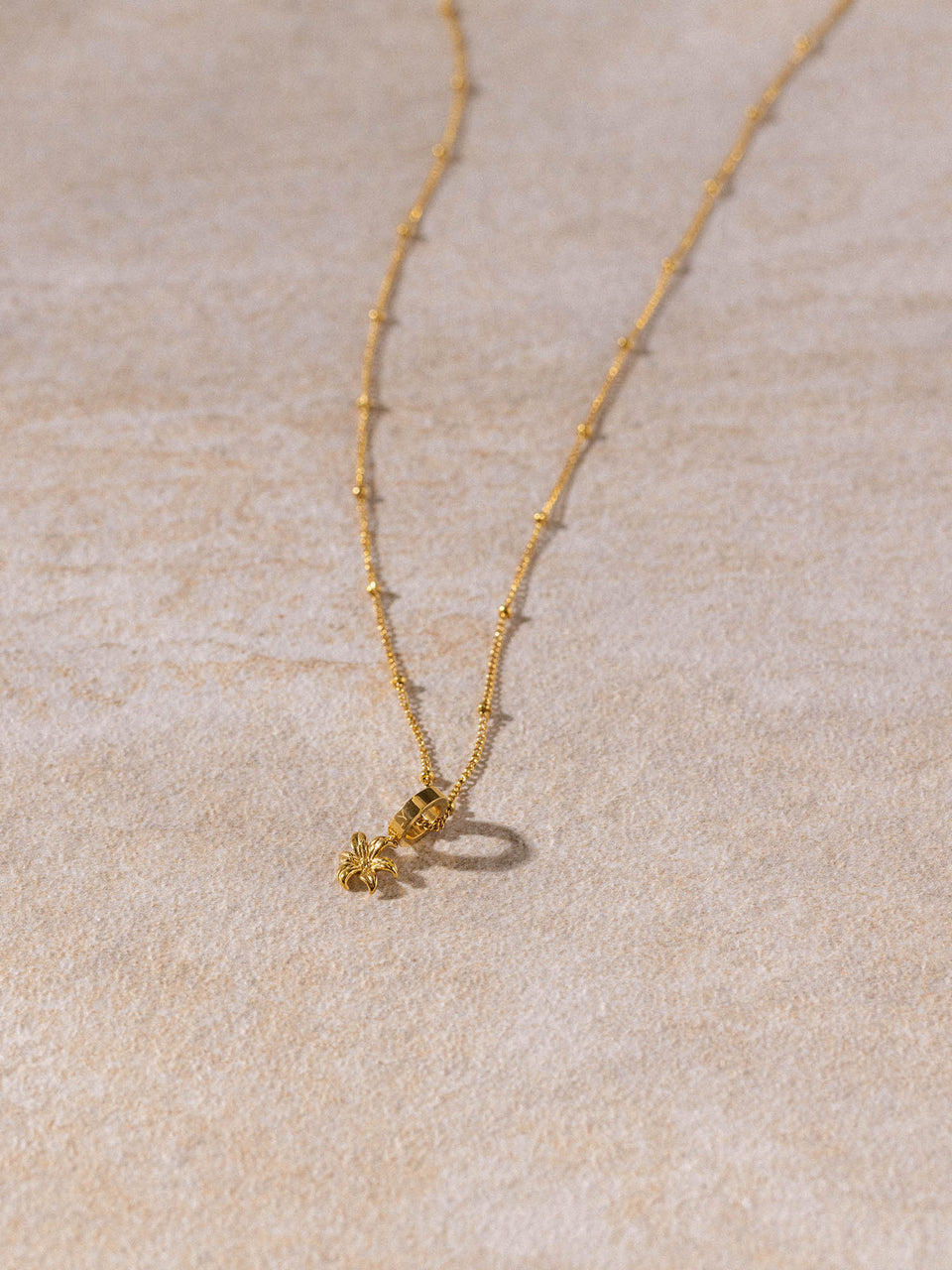 Feingliedrige Li’ili’i Kette aus 18K vergoldetem Edelstahl mit Blumenanhänger auf sandfarbenem Hintergrund PURELEI