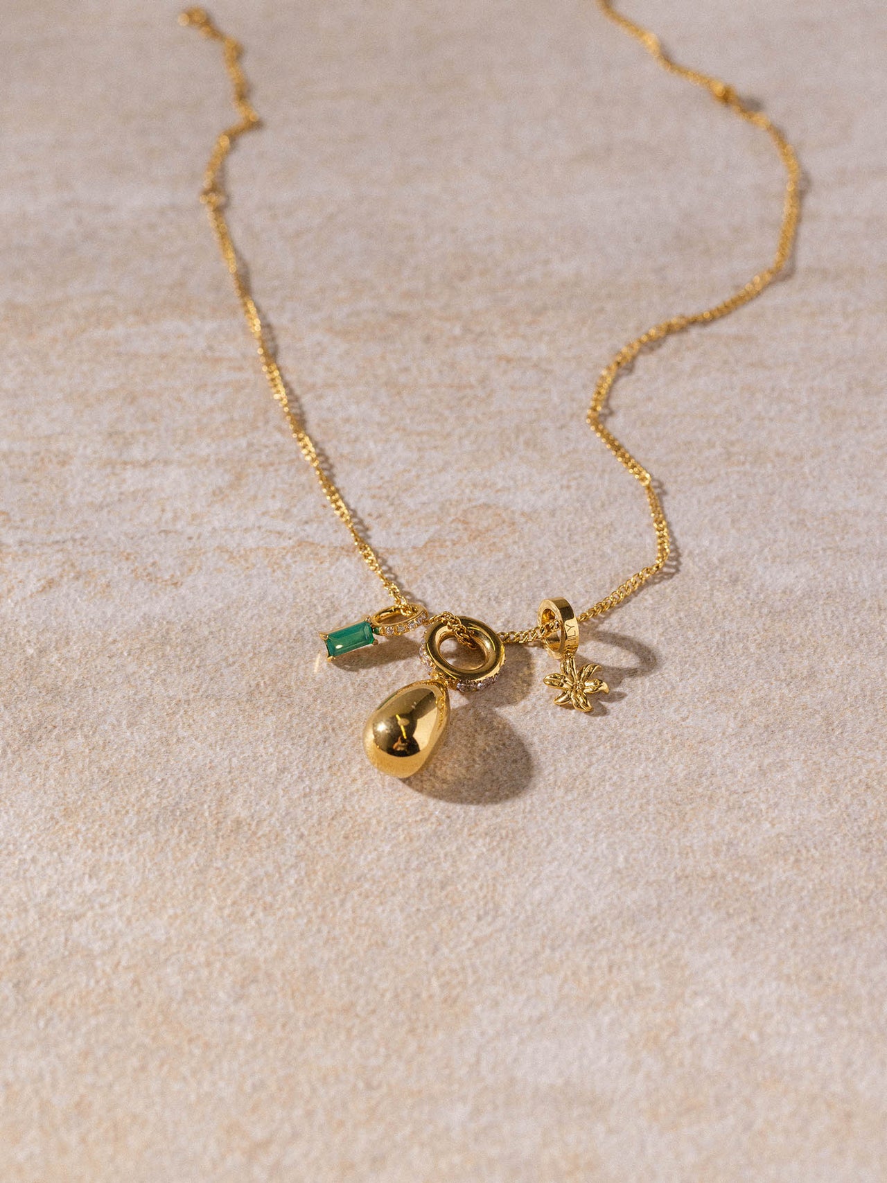 Feine PURELEI Kaula Kette aus 18K vergoldetem Edelstahl mit zarten Anhängern auf warmem Sand, elegant und feminin.