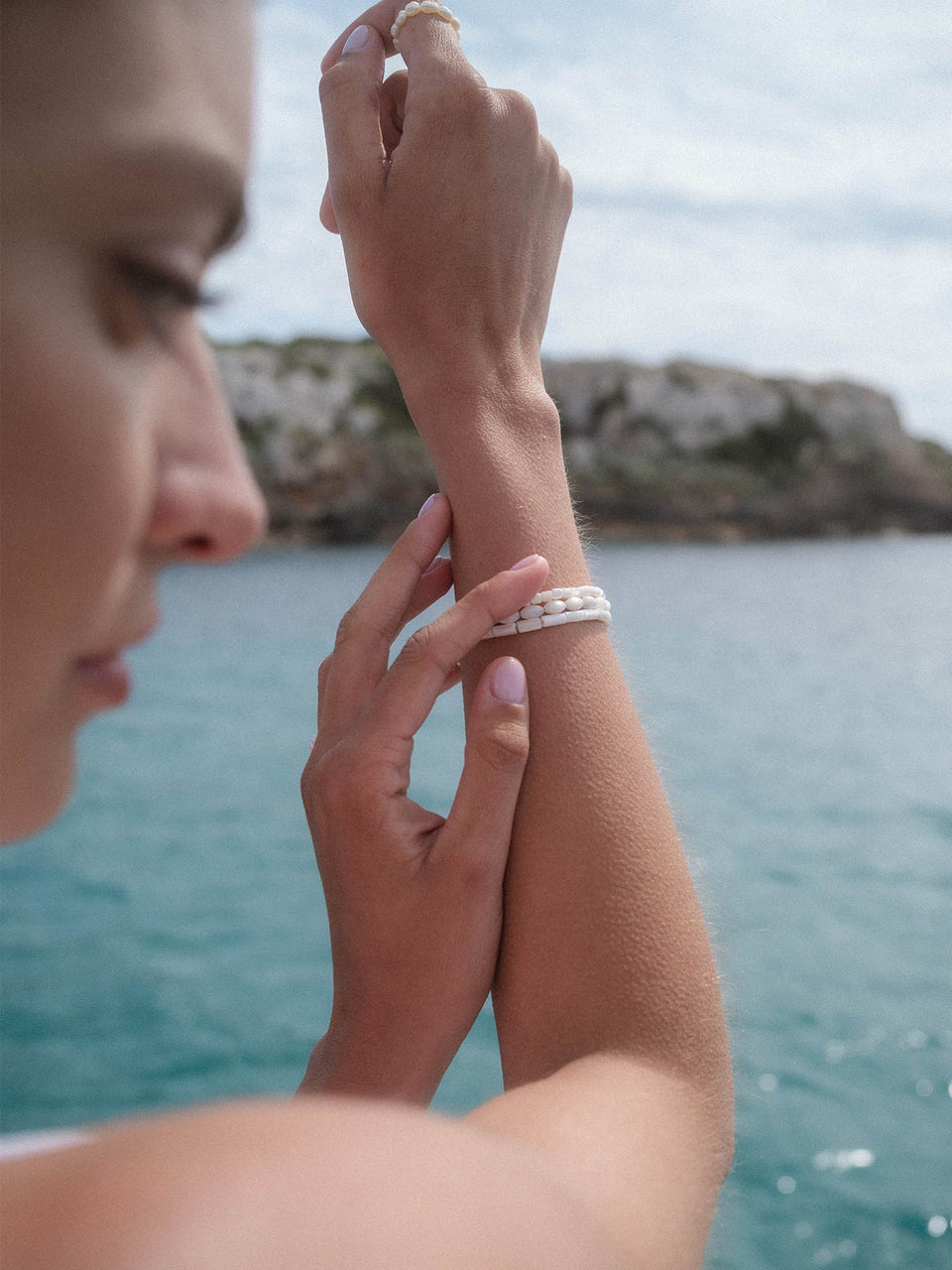 Summer Breeze Armband mit weißen Muschelperlen, goldfarben, PURELEI am Handgelenk vor Meereskulisse.