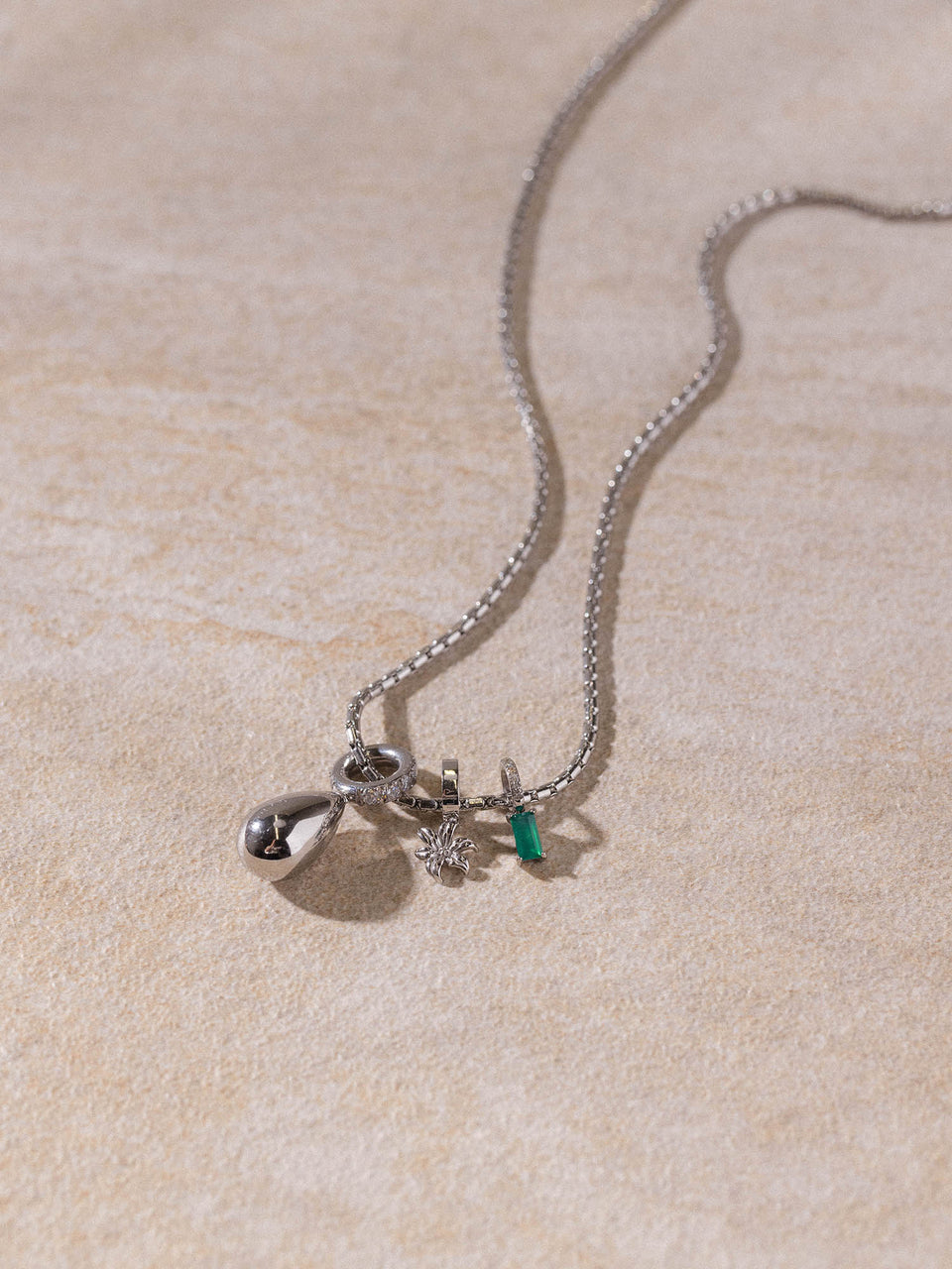 PURELEI Resonance Kette aus silberfarbenem Edelstahl mit Tropfen- und Blumenanhänger auf sandfarbenem Untergrund