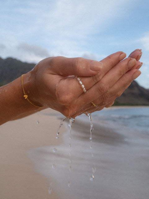 Zarte PURELEI Perlenarmbänder in Gold und Roségold, getragen am Strand mit Wassertröpfchen.