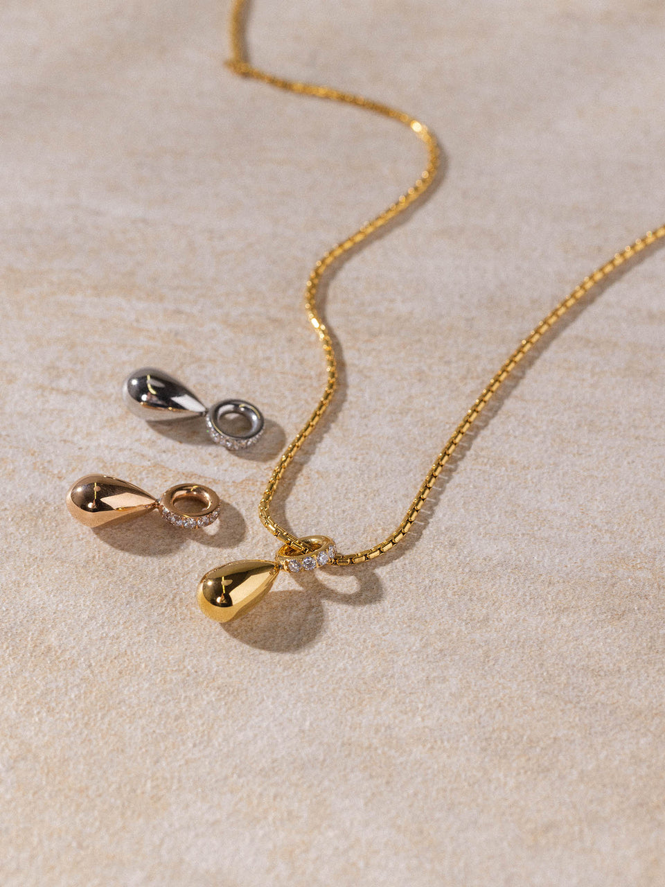 PURELEI Resonance Kette in Gold, Silber und Roségold mit tropfenförmigem Anhänger, clean & elegant auf sandfarbenem Hintergrund