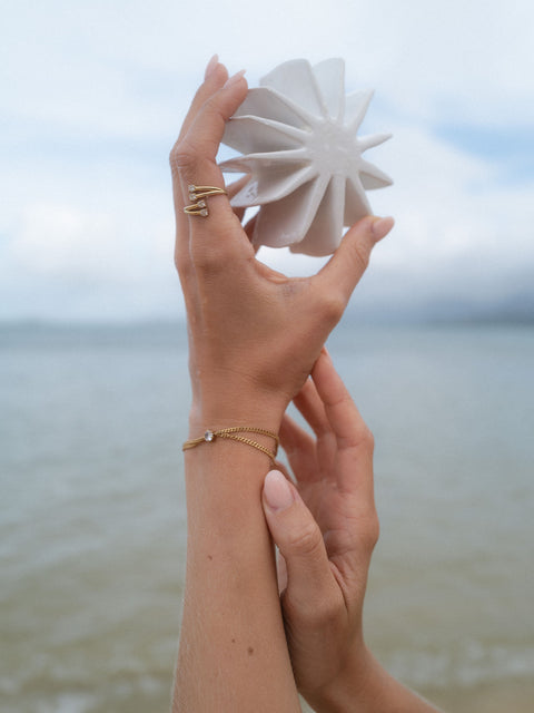 Zarte, roségoldene Armbänder und Ringe von PURELEI, getragen am Strand mit blühendem Hintergrund.