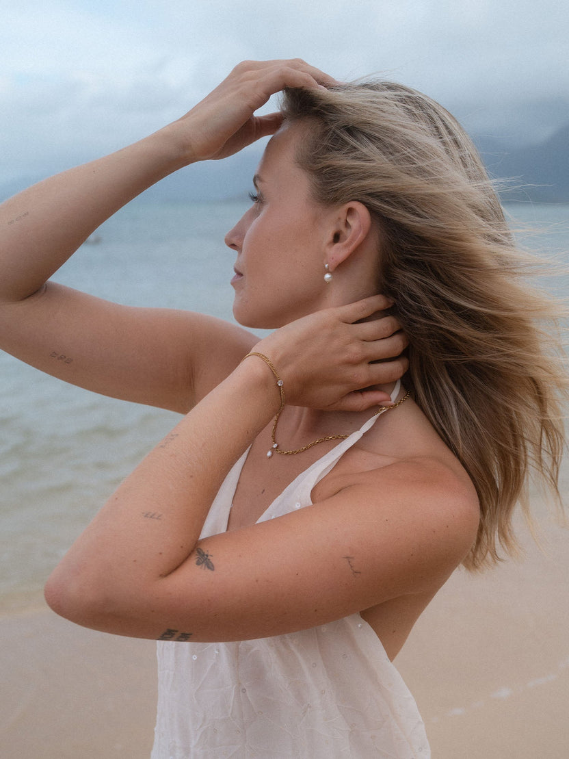 Elegante PURELEI Perlenohrringe und zartes Armband am Strand, umgeben von sanften Wellen.