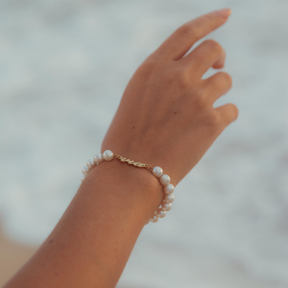 Via Pearl Perlenarmband von PURELEI mit goldenem via-Schriftzug und Muschelperlen am Strand getragen, elegant und feminin.
