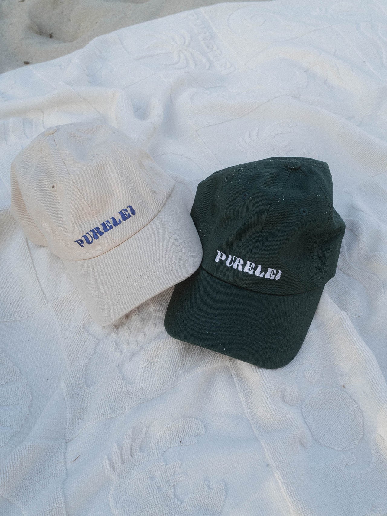 PURELEI Island Kappe aus Baumwolle in Beige und Blau, mit gesticktem Schriftzug, lässig am Strand inszeniert.