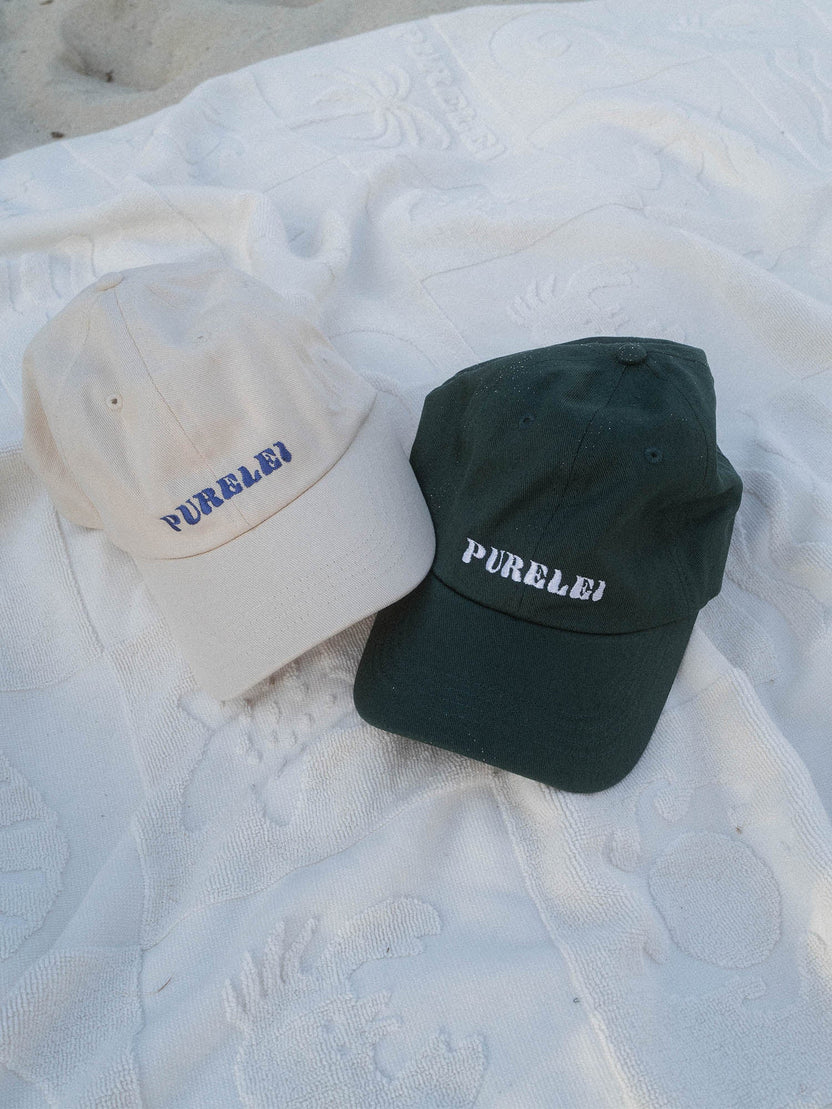 PURELEI Island Kappe aus Baumwolle in Beige und Blau, mit gesticktem Schriftzug, lässig am Strand inszeniert.