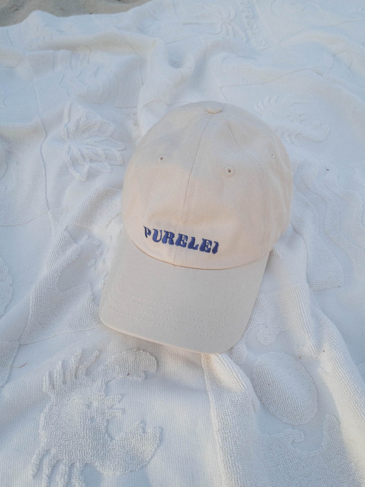 PURELEI Island Kappe aus beige Baumwolle mit blau gesticktem Schriftzug, elegant und lässig auf hellem Stoff.