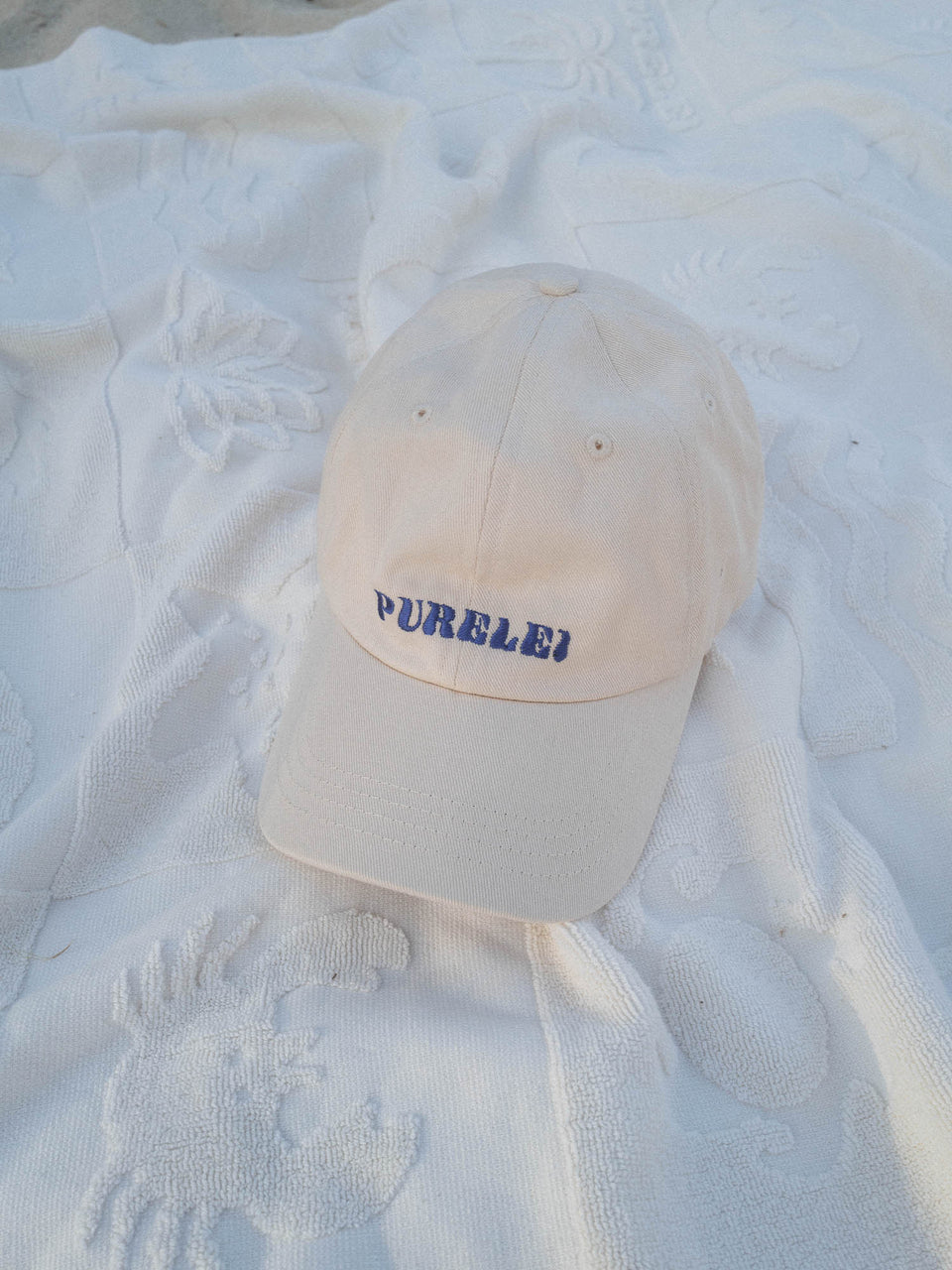 PURELEI Island Kappe aus beige Baumwolle mit blau gesticktem Schriftzug, elegant und lässig auf hellem Stoff.
