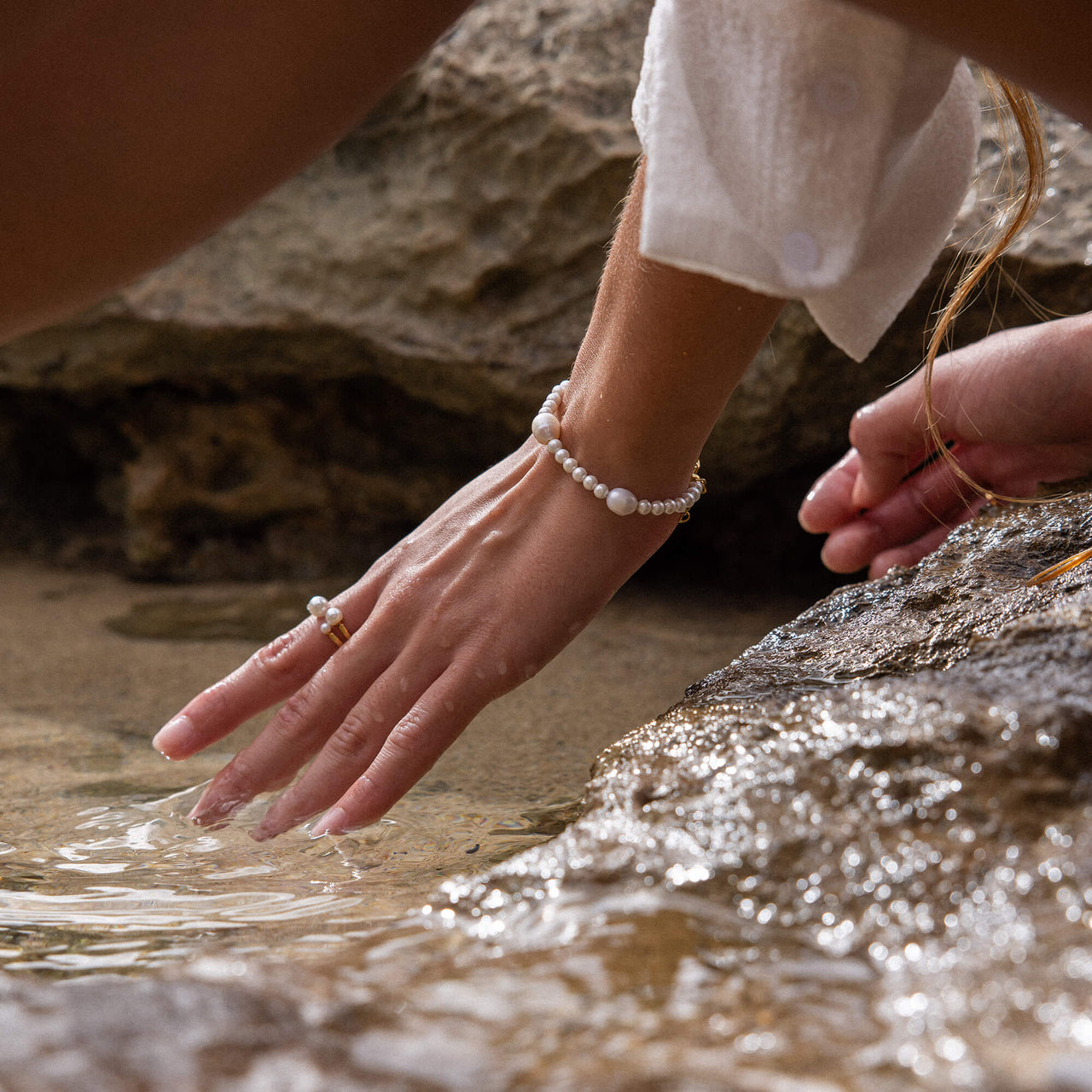 PURELEI Finesse Ring aus 18K Gold mit Muschelperlen, fein und feminin, getragen am Hand vor Naturstein und Wasser.