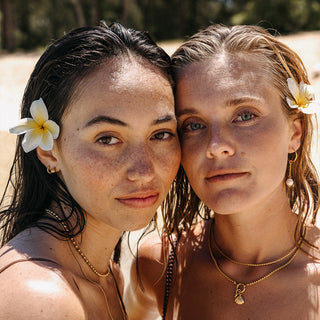 Zwei Frauen mit Schmuck von PURELEI, Muschel-Anhänger und Blumen im Haar, am Strand.