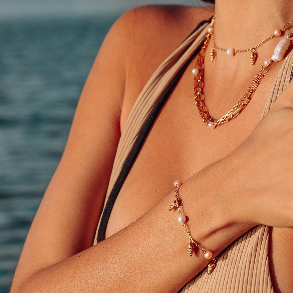 PURELEI Akala Armband aus vergoldetem Edelstahl mit Muschel- und Süßwasserperlen, getragen im Sonnenlicht am Meer