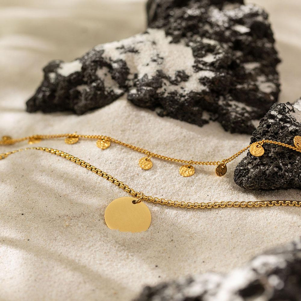 Olomana Ketten-Set von PURELEI, goldene Edelstahlketten mit Münzanhängern, elegant im Sand mit Lavasteinen präsentiert.