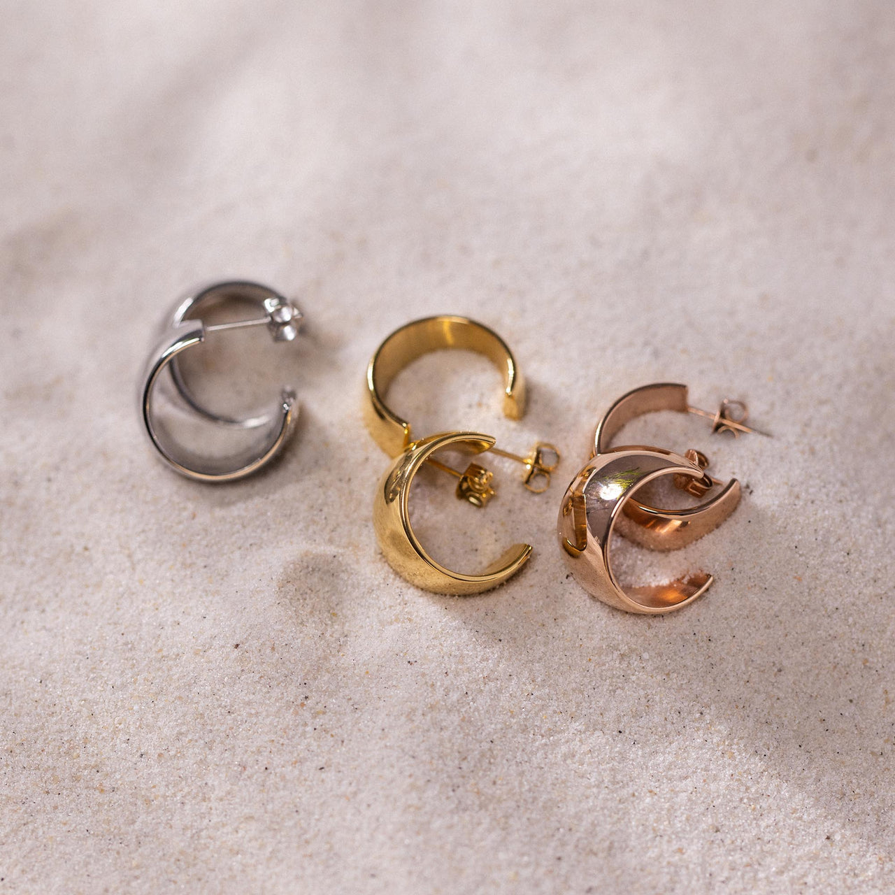 Bold Ohrringe aus gold, Silber und Roségold liegen elegant auf hellem Sand, pure feminine Ästhetik von PURELEI.